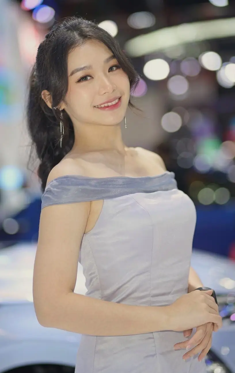 Brand ambassador bergaun perak off-shoulder tersenyum di area pameran otomotif