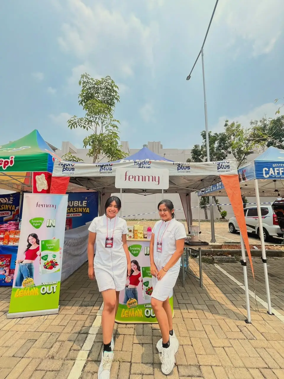 Two brand ambassadors in white uniforms in front of a product tent at an outdoor bazaar