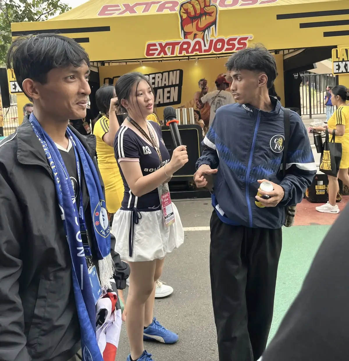 Seorang brand ambassador dengan mikrofon berbincang dengan suporter sepak bola di aktivasi brand