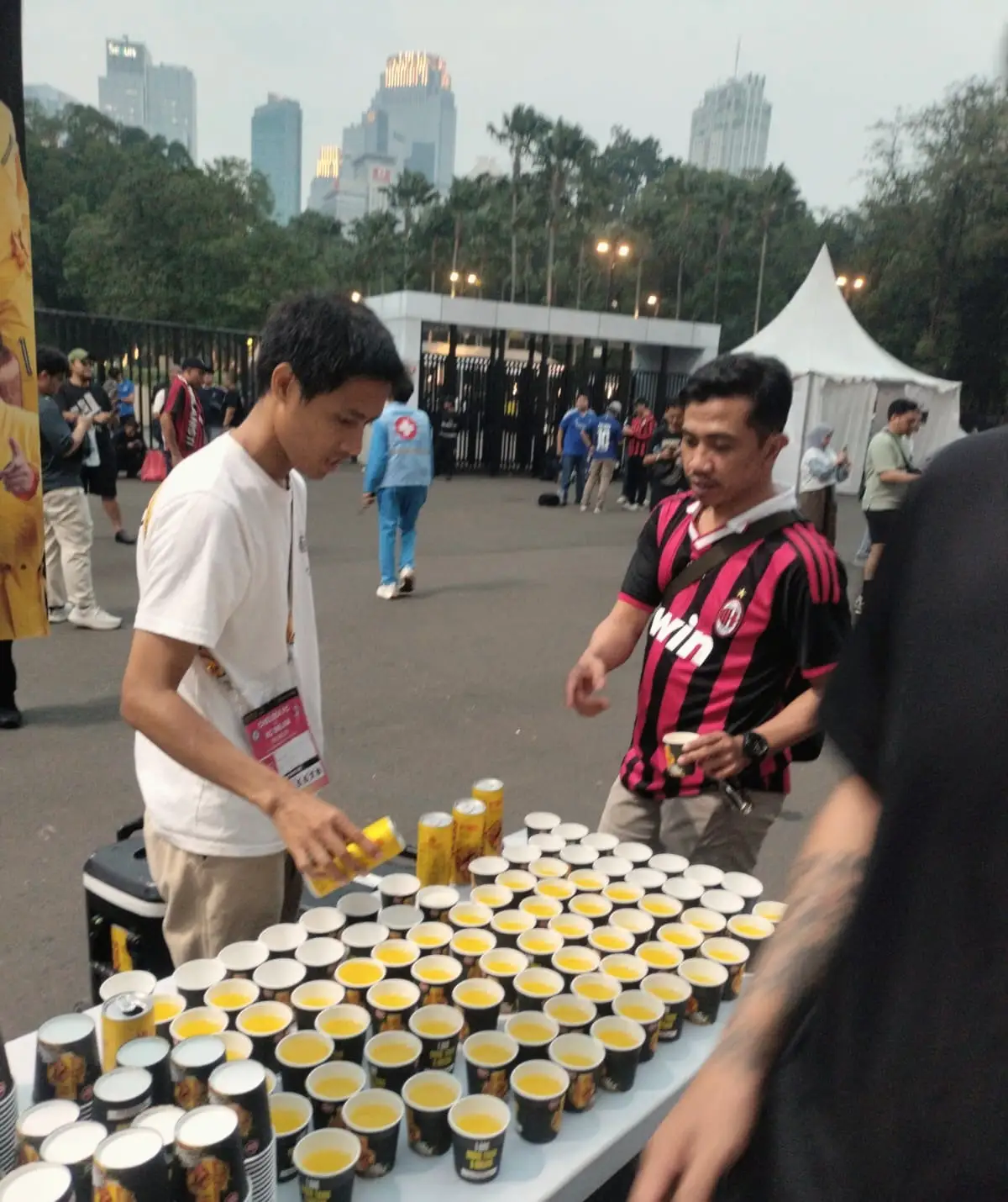 Kru menuang deretan sampel minuman di meja pada acara luar ruang, dengan latar gedung kota