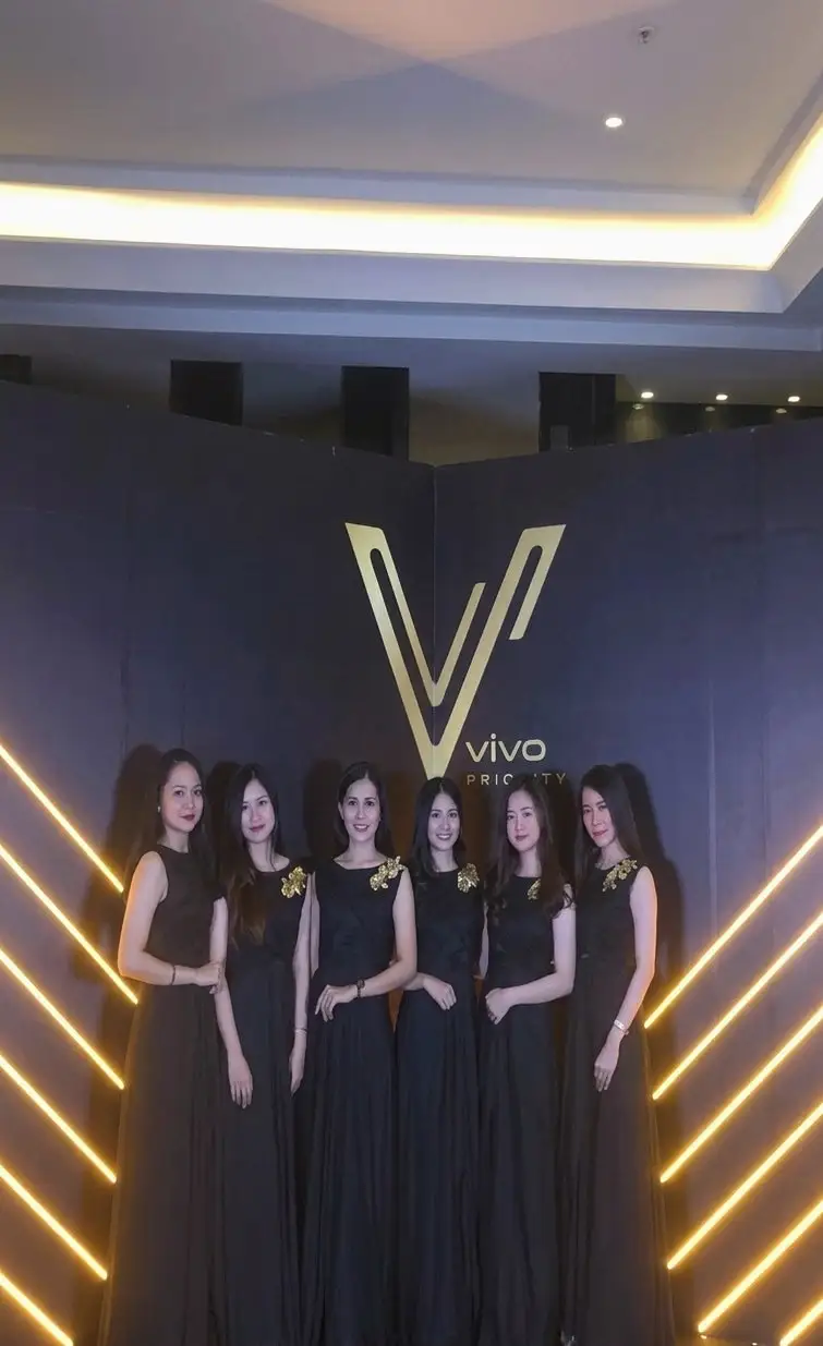 Enam brand ambassador bergaun hitam berbaris di bawah logo emas pada peluncuran smartphone